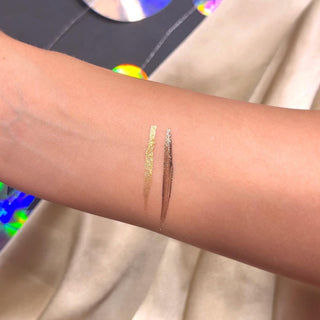 Close-up of a person's arm with two P.Louise Glisten To Your Heart Glitter Eyeliner Dusk Diva Set gold and brown swatches on against a blurred background
