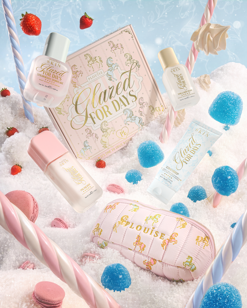 A styled flat-lay of a skincare gift set featuring a pink box labeled 'Glazed for Days' and multiple matching pink and pastel products including bottles, tubes, and a zip pouch with a carousel motif, arranged in white faux snow with candy props and strawberries.
