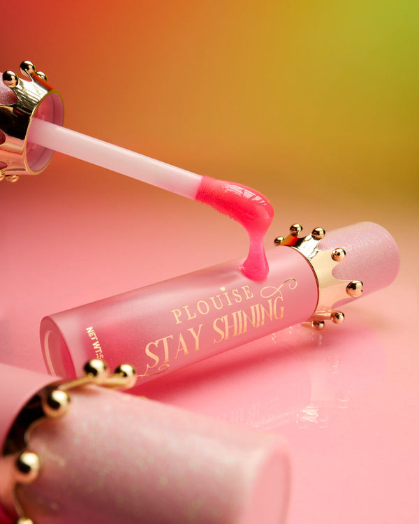 Pink tube of P.Louise STay SHining glossy lip oil laying on a pink surface with a lip oil wand resting against the tube showing the product