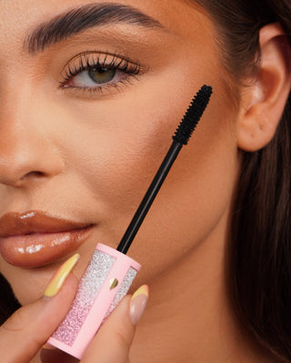 Close-up of a P.Louise Cry Baby Mascara wand for sensitive eyes held near a person's eye with a pink glittery mascara tube.