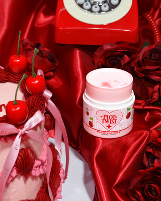 A container of P.Louise Plot Twist Makeup Remover Balm displayed on a red fabric surface with a decorative cherries and ribbon, alongside a vintage red phone and a pink and red cake.